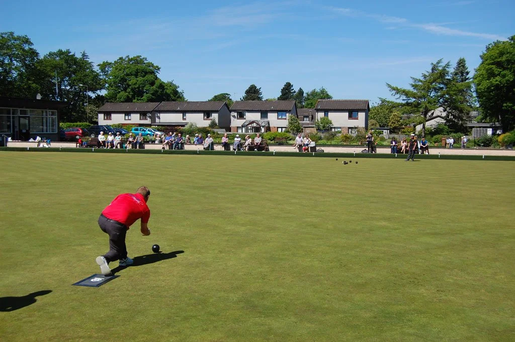 A man in a red shirt playing lawn bowls on a green field surrounded by spectators and houses in the background under a clear blue sky.
