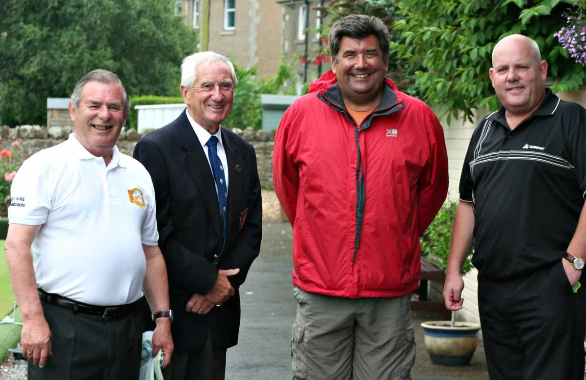 Four men standing outdoors in front of trees and a building, smiling at the camera.