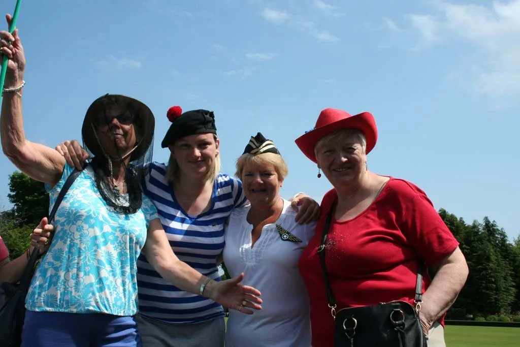 Four women standing outside under a blue sky, dressed in patriotic and vintage outfits, smiling and posing together.