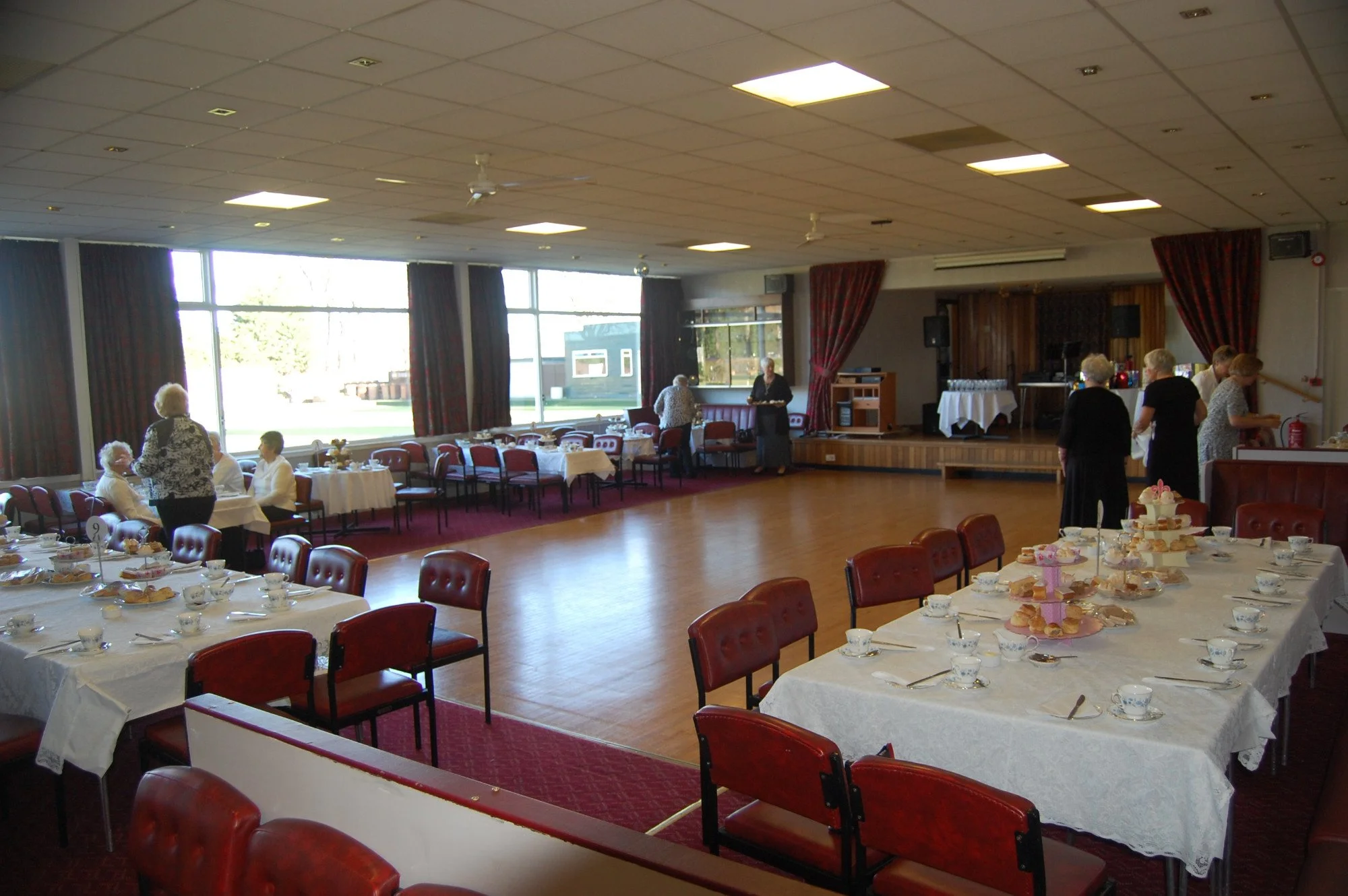 A banquet hall set up for an event with round tables covered in white tablecloths, decorated with tea cups and tiered serving trays with desserts. People are mingling and preparing in the background near a small stage, with large windows and red curt