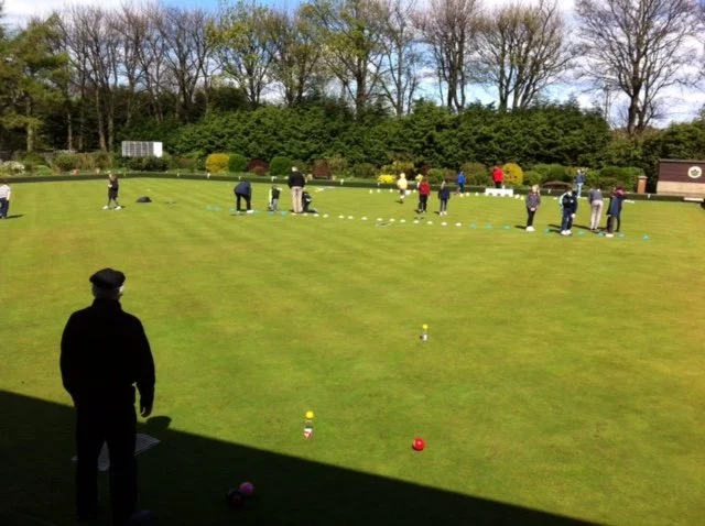Group of people practicing lawn bowls on a green field outdoors, with some individuals bending down to aim their bowls, surrounded by trees and clear skies.
