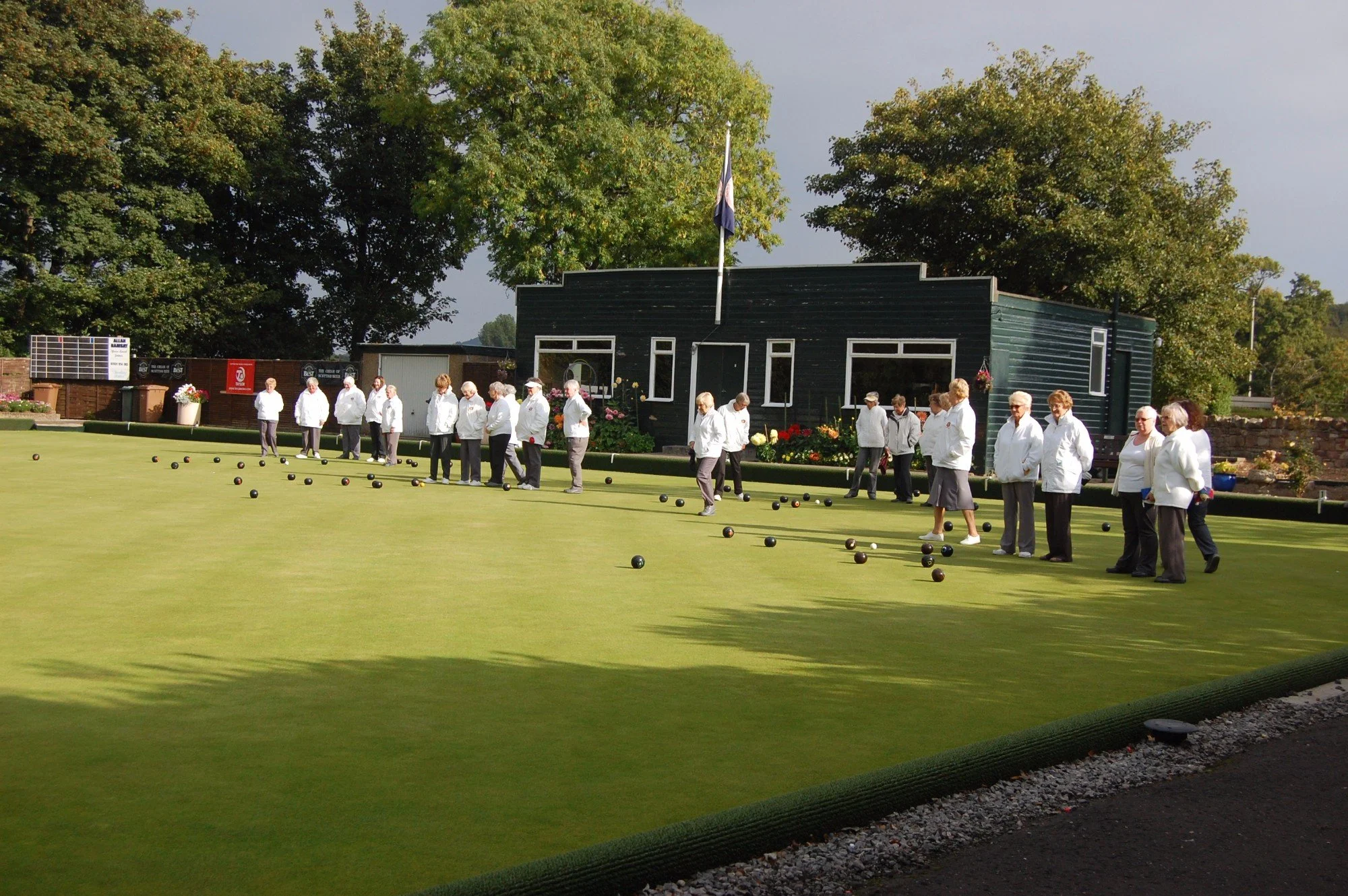 People gathered on a lawn playing bocce, with a dark green building and large trees in the background under cloudy skies.