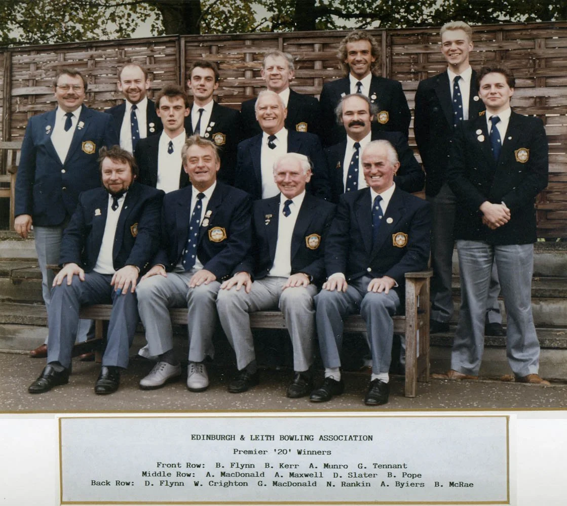 Group photo of men in formal suits with badges, posed in front of a wooden fence, celebrating Edinburgh & Leith Bowling Association Premier '20' winners.