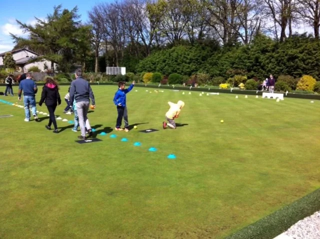 Children participating in a golf training session on a golf green during daytime, with children kneeling and others walking in line, surrounded by trees and bushes.