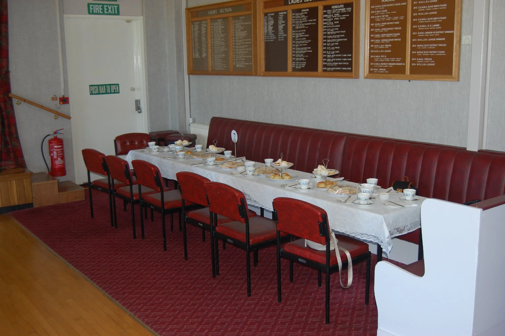 A long table set for afternoon tea with cups, saucers, and plates with scones and sweets, in a room with a red carpet, red cushioned chairs, a wall-mounted menu board, and a fire exit sign.