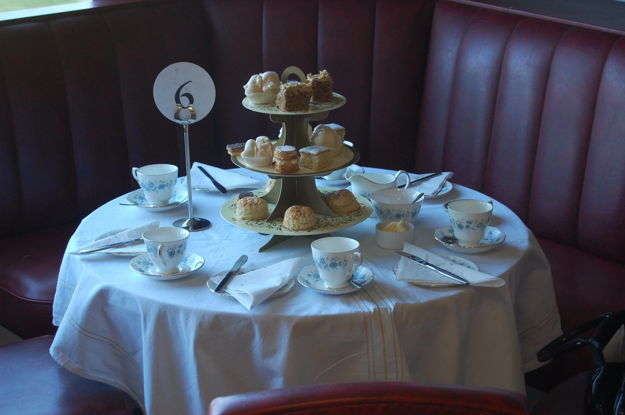 Table set for afternoon tea with a three-tier stand filled with assorted cakes, scones, and desserts, surrounded by teacups, saucers, and utensils, in a cozy dining environment with red upholstered seating.
