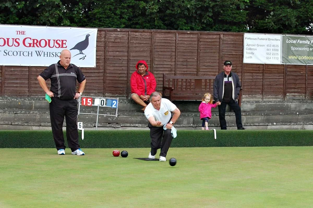A man in a white shirt bowls on a green lawn during a lawn bowling game, with spectators, including a child and an older man, sitting and standing behind. There are scoreboards and banners in the background.