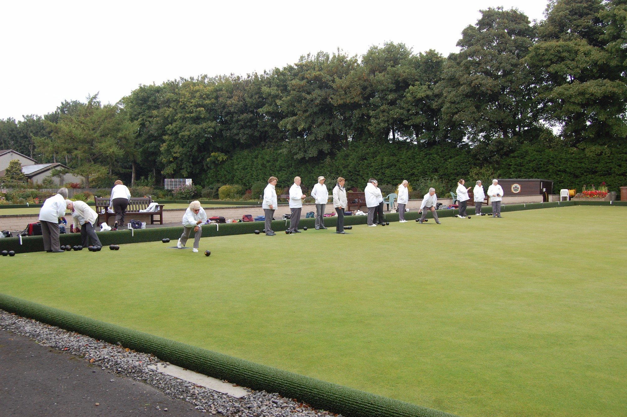 Group of elderly people playing lawn bowls outdoors, lined up and approaching bowls on a well-maintained green, with trees and garden area in the background.