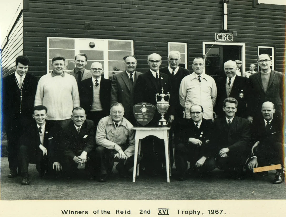 A group of 14 men posing outside a building with dark siding, some wearing ties and suits, some in uniforms, with a large trophy and a plaque on a table in front of them, celebrating their win of the Reid 2nd XVI Trophy in 1967.
