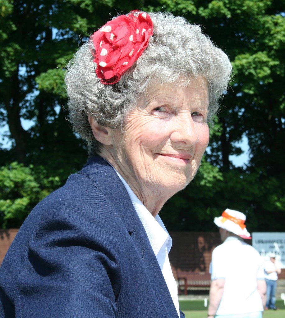 A smiling elderly woman with curly gray hair wearing a red headband with white polka dots, dressed in a dark navy blazer and white shirt, outdoors with green trees in the background and another person wearing a large white hat with orange accents.
