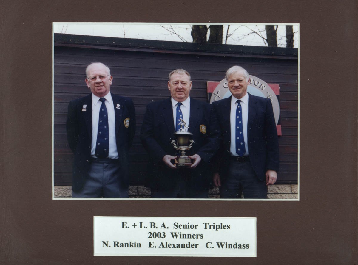 Three men dressed in dark suits and ties standing outside in front of a wooden wall. The man in the middle holds a trophy. A sign with the text "Loyalty Culton" is partially visible on the wall behind them. The photo has a brown mat with a white labe
