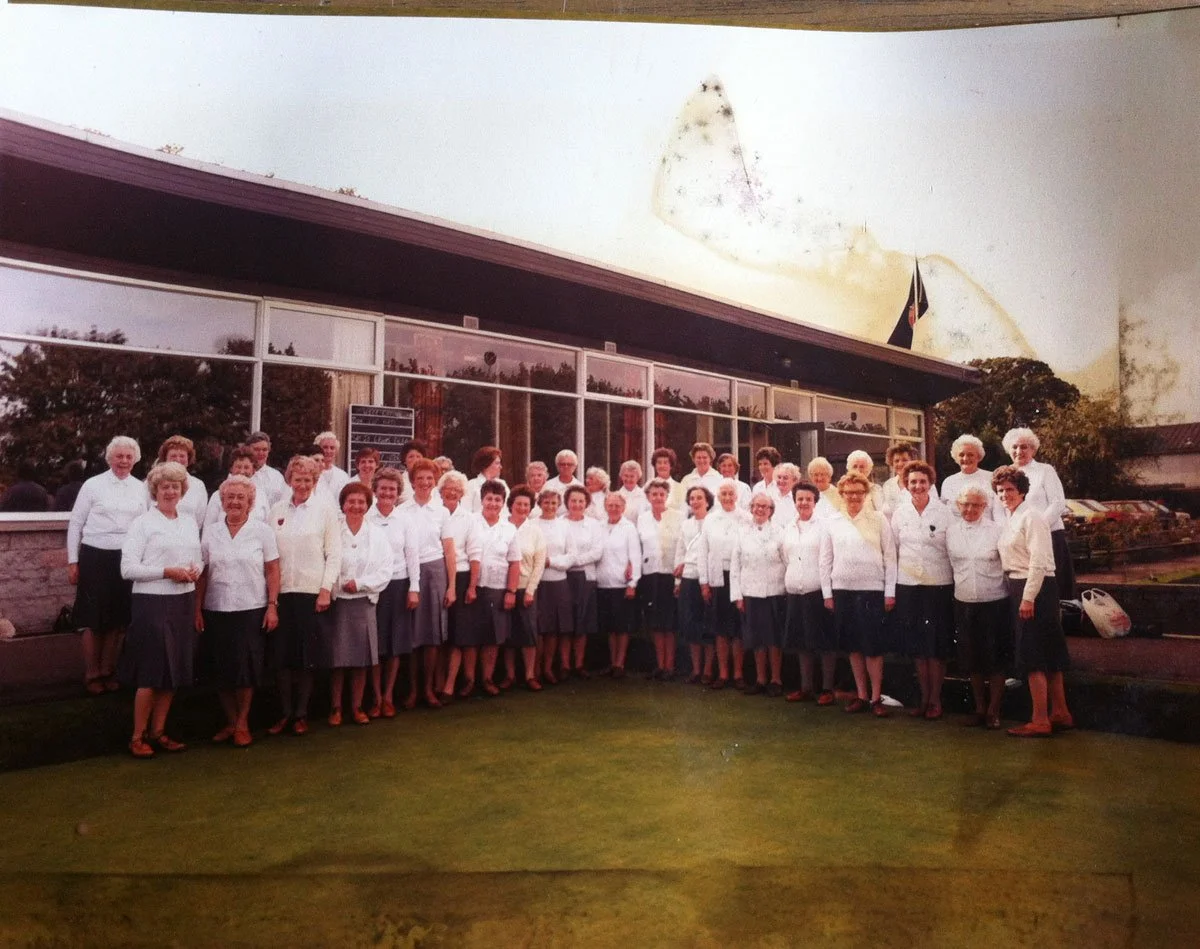 A group of women dressed in white blouses and dark skirts, standing outside a large building with glass windows, possibly a community or church building, during daytime.