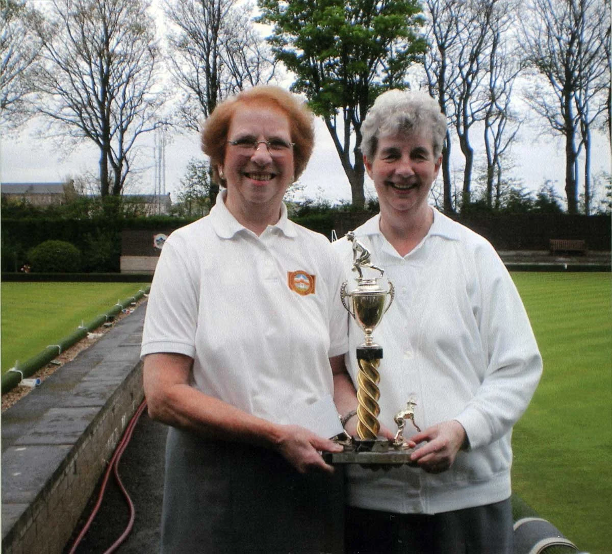 Two women in white shirts standing outdoors on a lawn, smiling, holding a large trophy, with trees and a bench in the background.