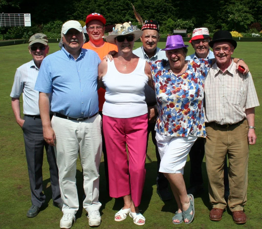 Group of eight older adults standing together on a golf course, smiling, with greenery in the background. They are dressed casually, some wearing hats and sunglasses.