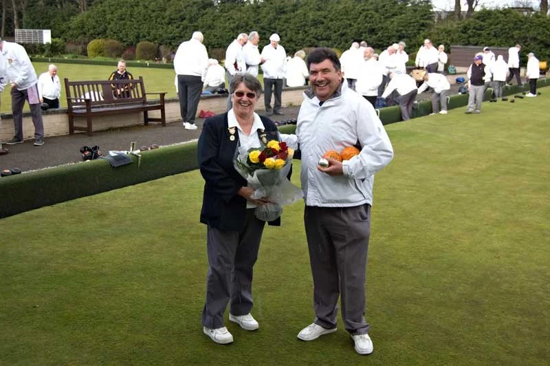 People playing lawn bowls on a green field with spectators and officials watching in the background. Two individuals in the foreground holding a bouquet of flowers and a trophy or prize.
