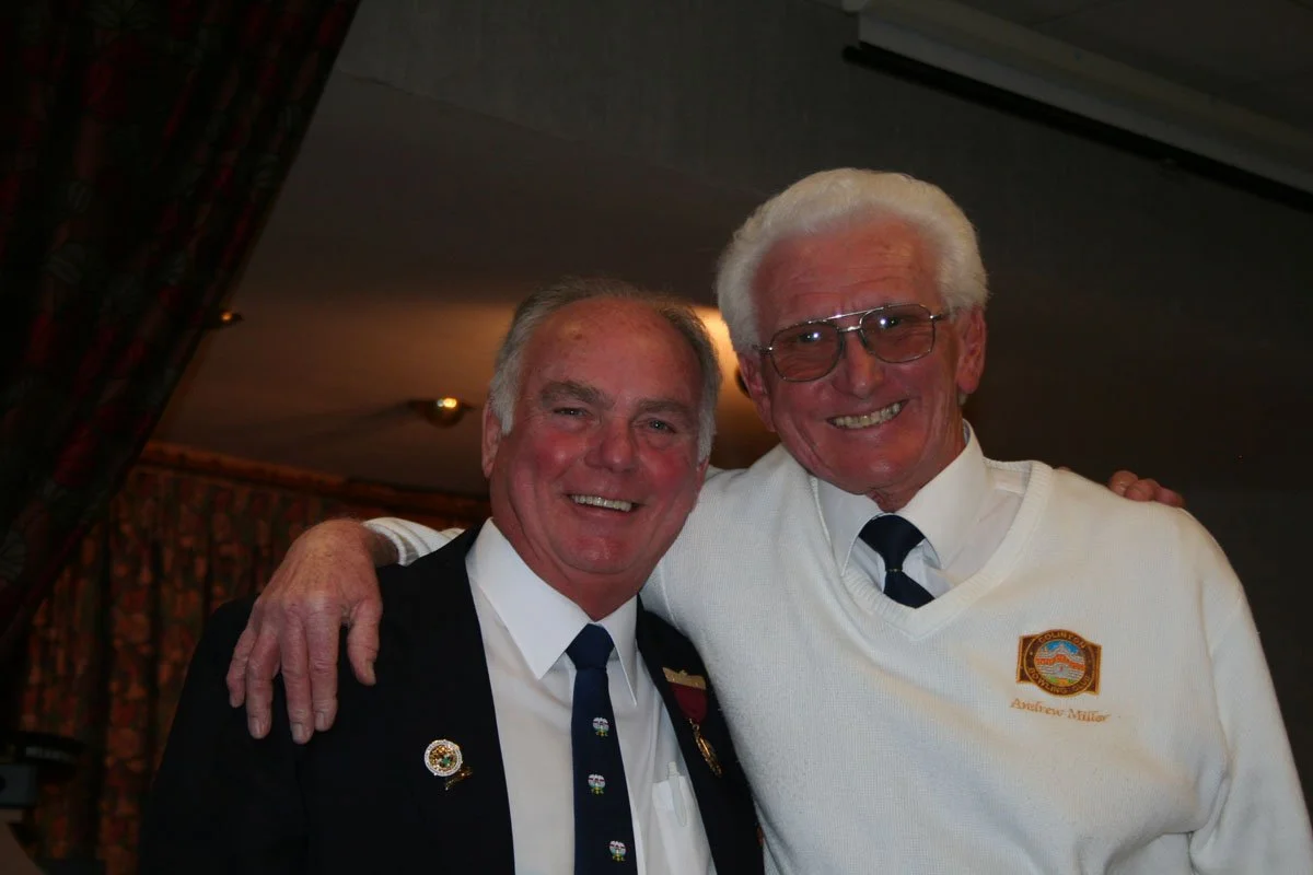 Two elderly men smiling and hugging, dressed in formal shirts and ties, indoors with curtain and ceiling lights in the background.