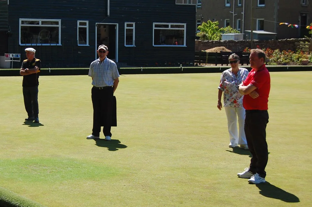 Three men and one woman standing on a golf green, with one man appearing to be preparing for a shot, and others watching on, in an outdoor setting on a sunny day.