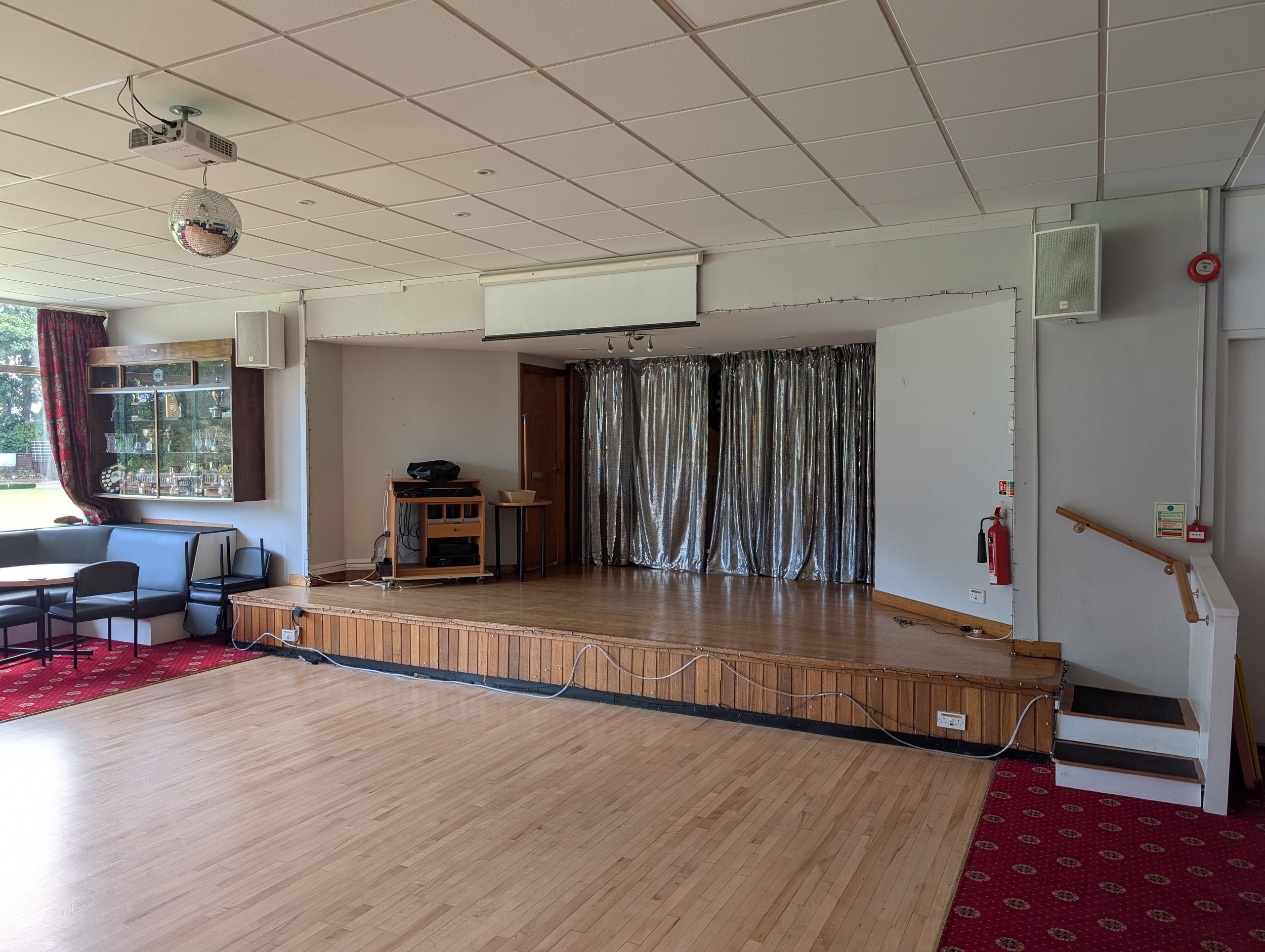 An empty community hall or event space with a stage, gold curtains, chairs, a disco ball hanging from the ceiling, and a window with a red patterned curtain on the left.