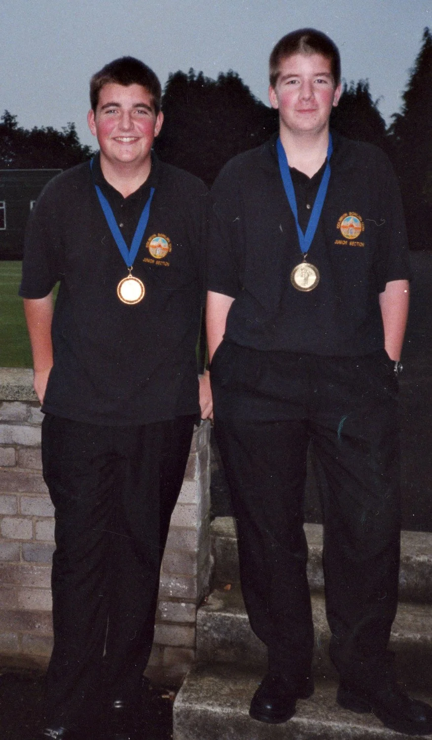 Two young men standing outdoors at dusk, both with medals around their necks, wearing dark uniforms with a logo on the chest, smiling at the camera.