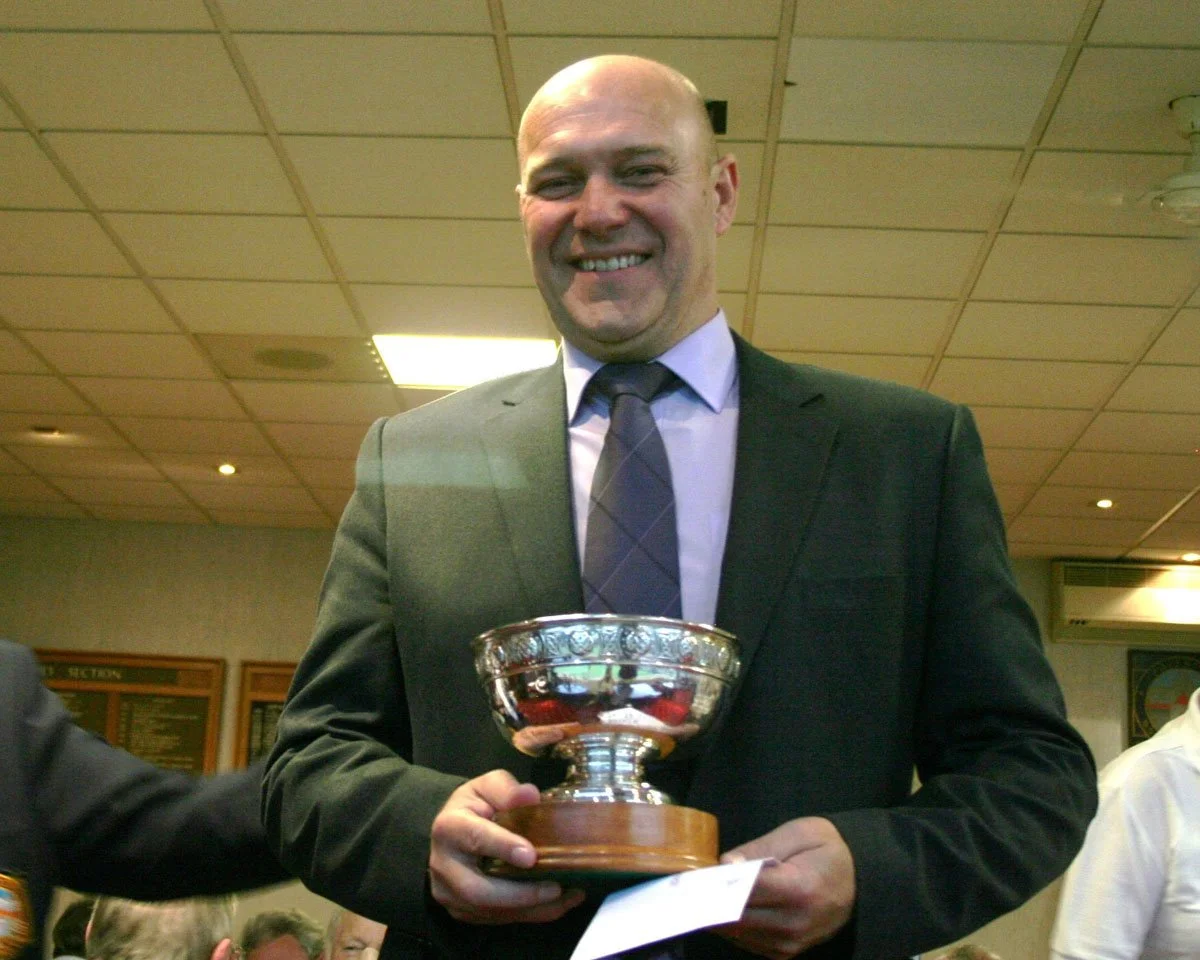 A man in a suit holding a trophy, smiling indoors.