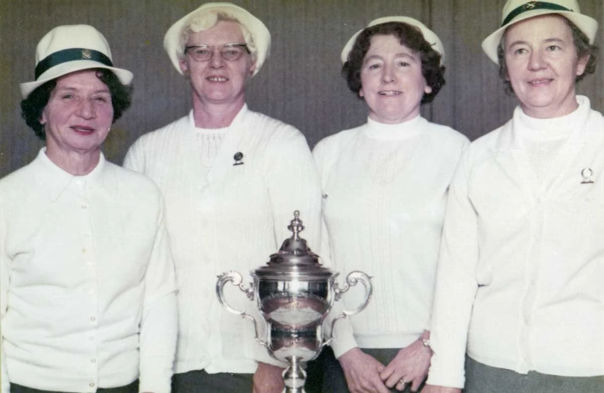 Four women and one man wearing white clothing and hats, standing behind a large trophy cup.