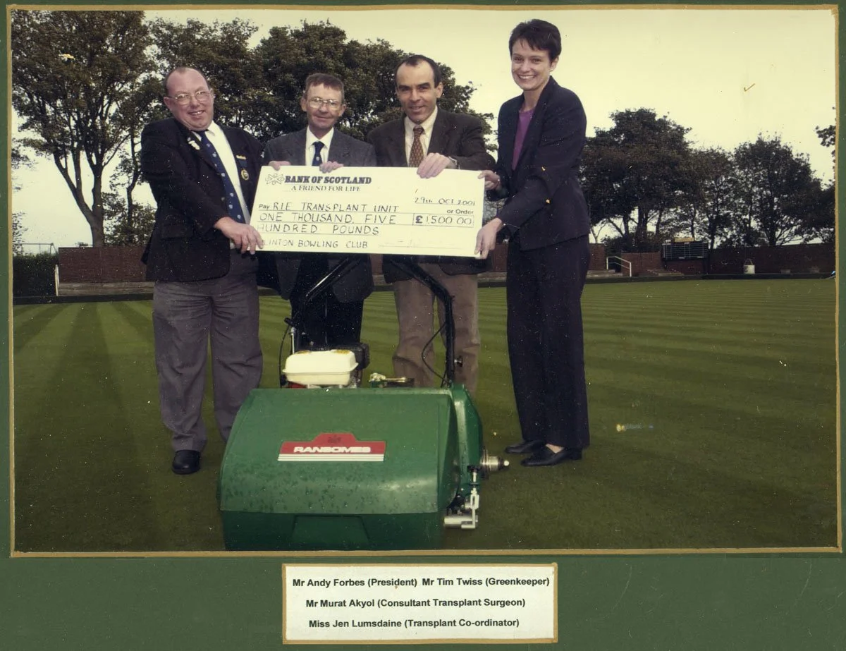 Four people standing on a football field holding a large check for £1,500 made out to the 'Rifle Transplant Unit'. The check is from the Bank of Scotland. The group includes Mr. Andy Forbes (President), Mr. Tim Twiss (Greenkeeper), Mr. Murat Akyol (C