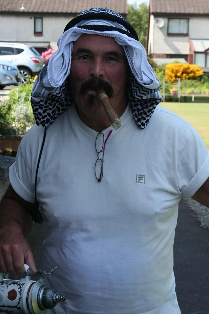 Man with a towel worn on his head, smoking a cigar, holding a large paint sprayer, standing outdoors with houses and cars in the background.