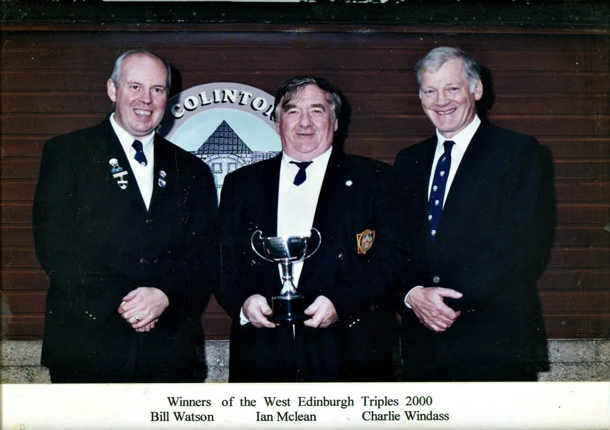 Three men dressed in formal black suits and ties, standing in front of a wooden wall with a sign that reads 'Collinton.' The man in the middle is holding a trophy. The caption below identifies them as winners of the West Edinburgh Triples in 2000: Bi