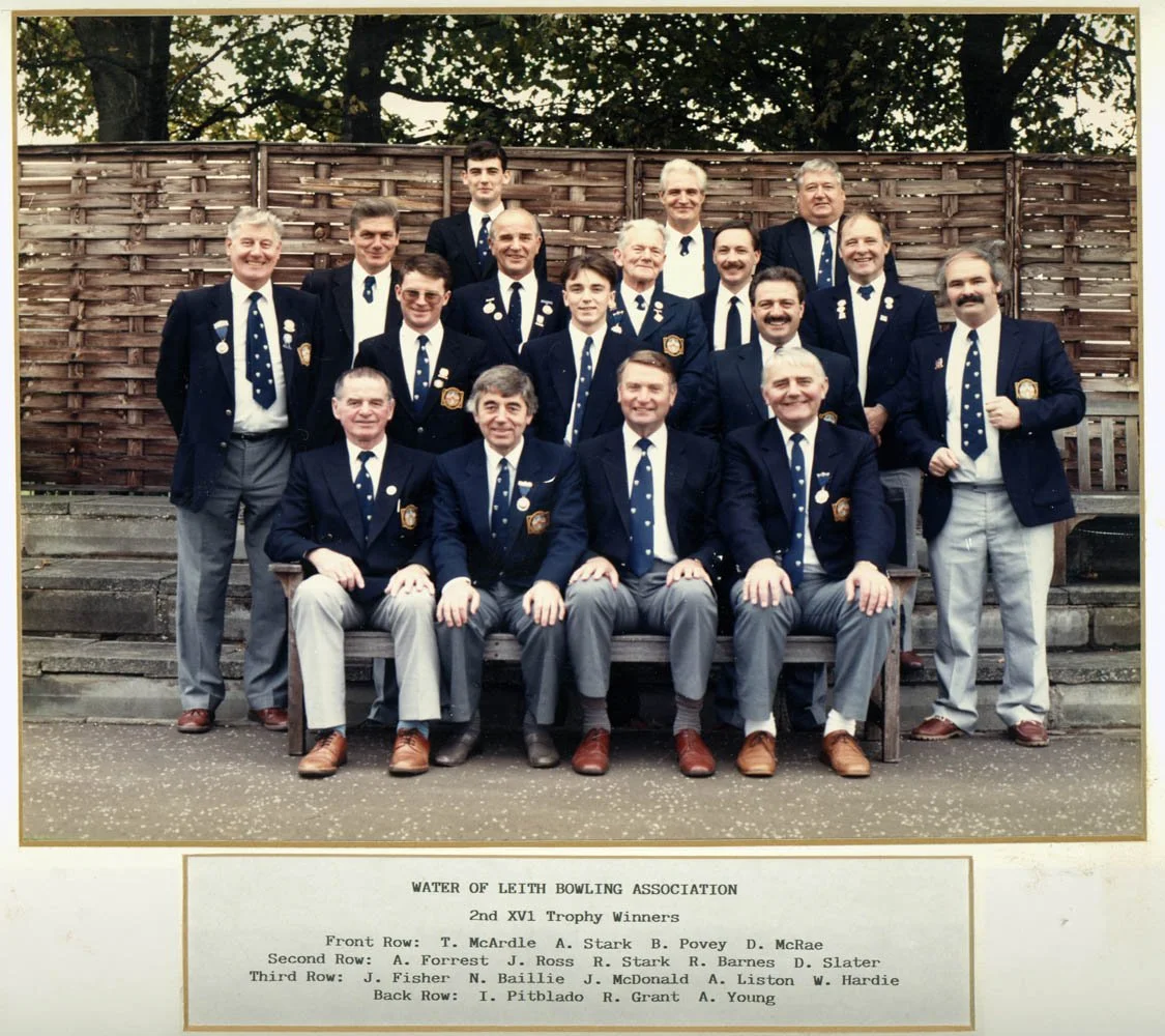 Group photo of men in navy blazers with pins, sitting and standing on stairs outdoors, celebrating Water of Leith Bowling Association 2nd XVI Trophy winners.