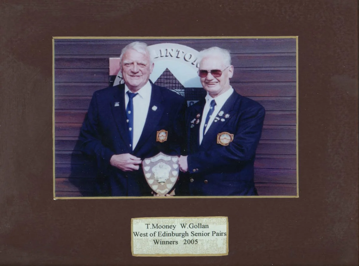 Two senior men in dark suits with medals and pins, standing in front of a wooden wall, holding a shield-shaped award plaque. The sign below reads: "T. Mooney W. Gollan West of Edinburgh Senior Pairs Winners 2005."