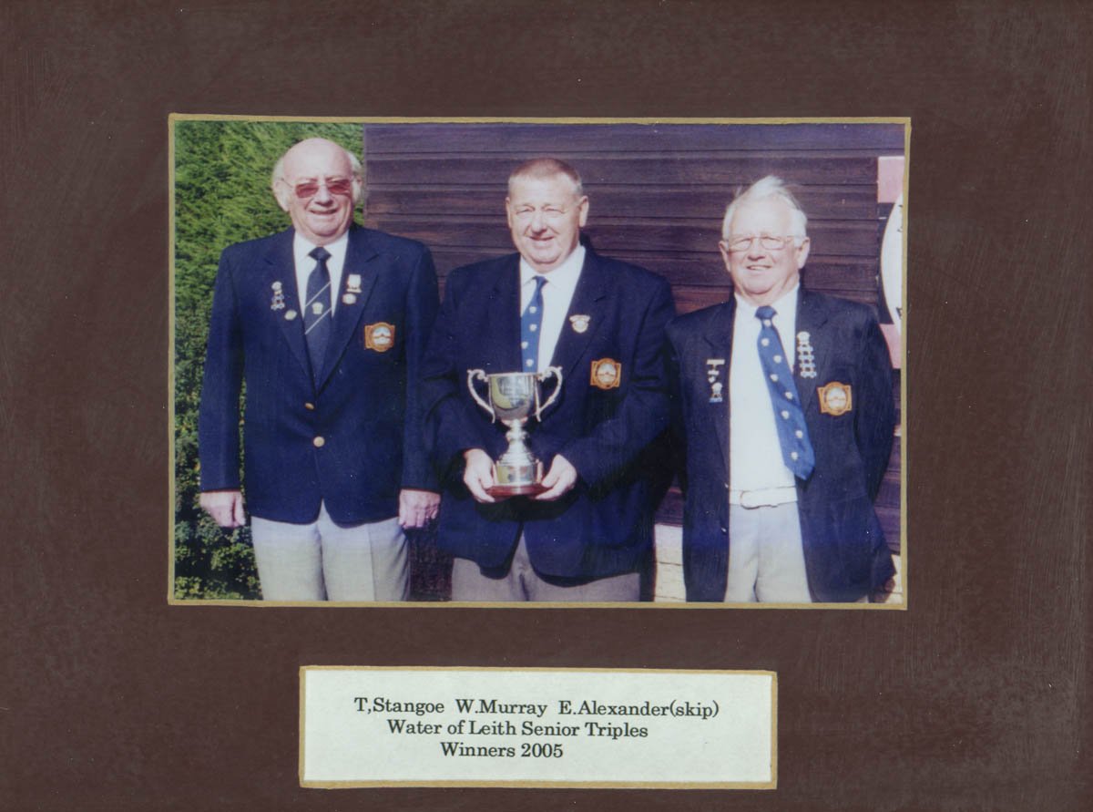 A framed photo of three elderly men in dark blazers wearing badges, standing outdoors in front of a building. The man in the middle is holding a silver trophy. A plaque below the photo reads: "T. Stangoe W. Murray E. Alexander (skip) Water of Leith S