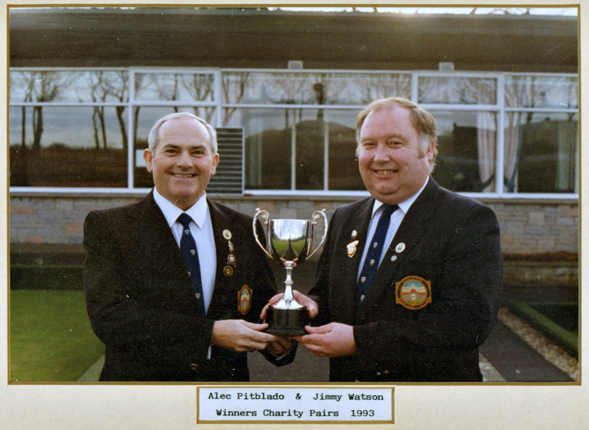 Two men in suits holding a trophy. One man is named Alec Pitblado and the other Jimmy Watson, both winners of charity pairs in 1993. They are standing outside in front of a building with large windows and trees reflected in the glass.