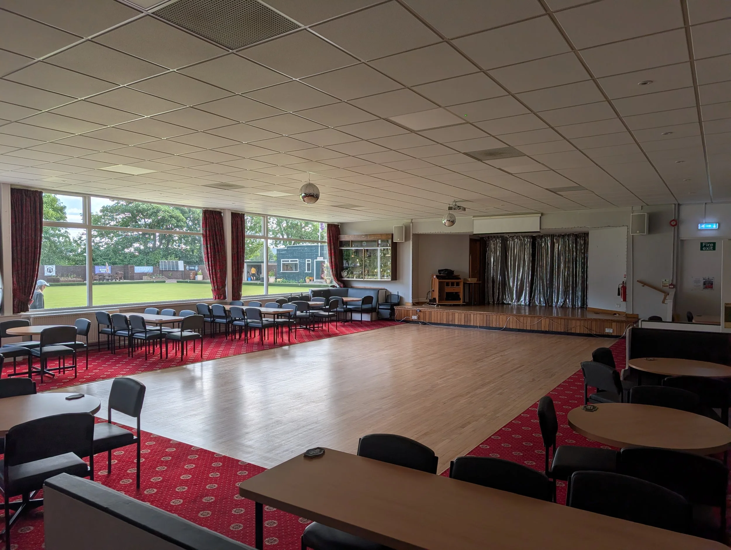 Empty event hall with large windows, red carpet, chairs, and tables, staged area with curtains, ceiling with lighting and speakers, and a view of a green field outside.