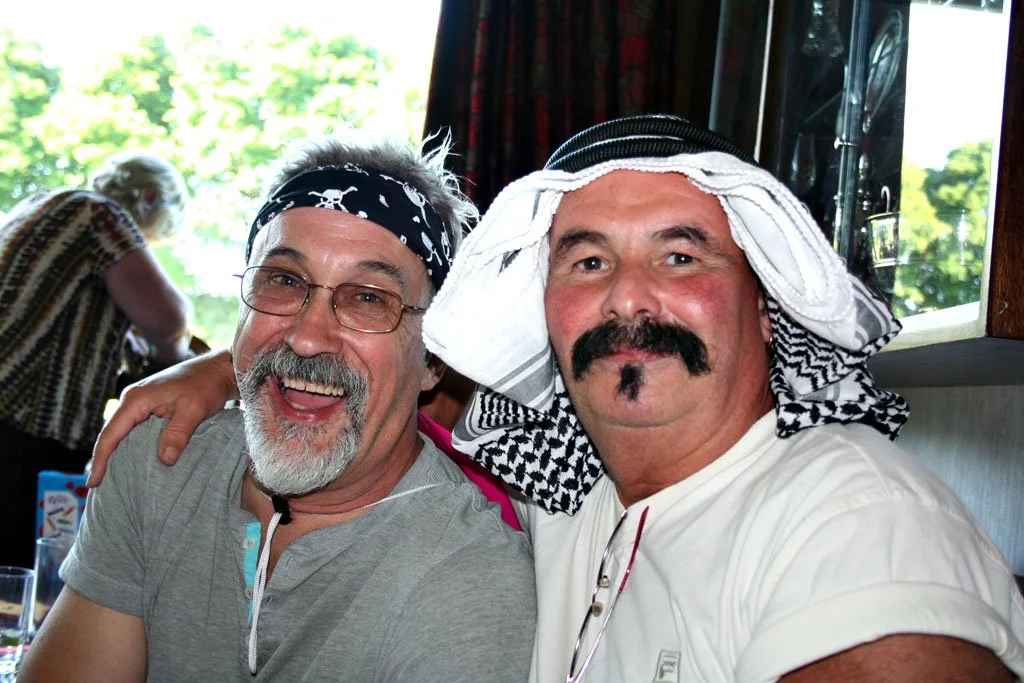Two men smiling and posing together indoors, one with a black bandana and glasses, the other with a traditional Middle Eastern head covering and a mustache, with a woman in a striped shirt in the background.