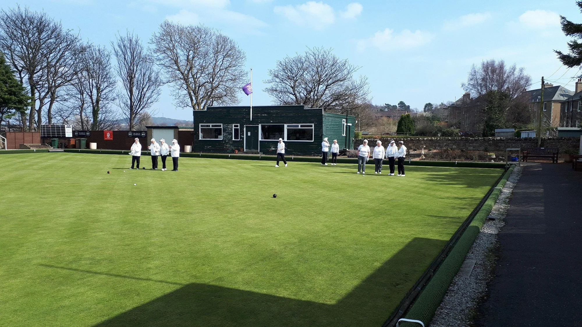 Senior citizens playing lawn bowls on a green field under a blue sky with scattered clouds, surrounded by trees and residential buildings.