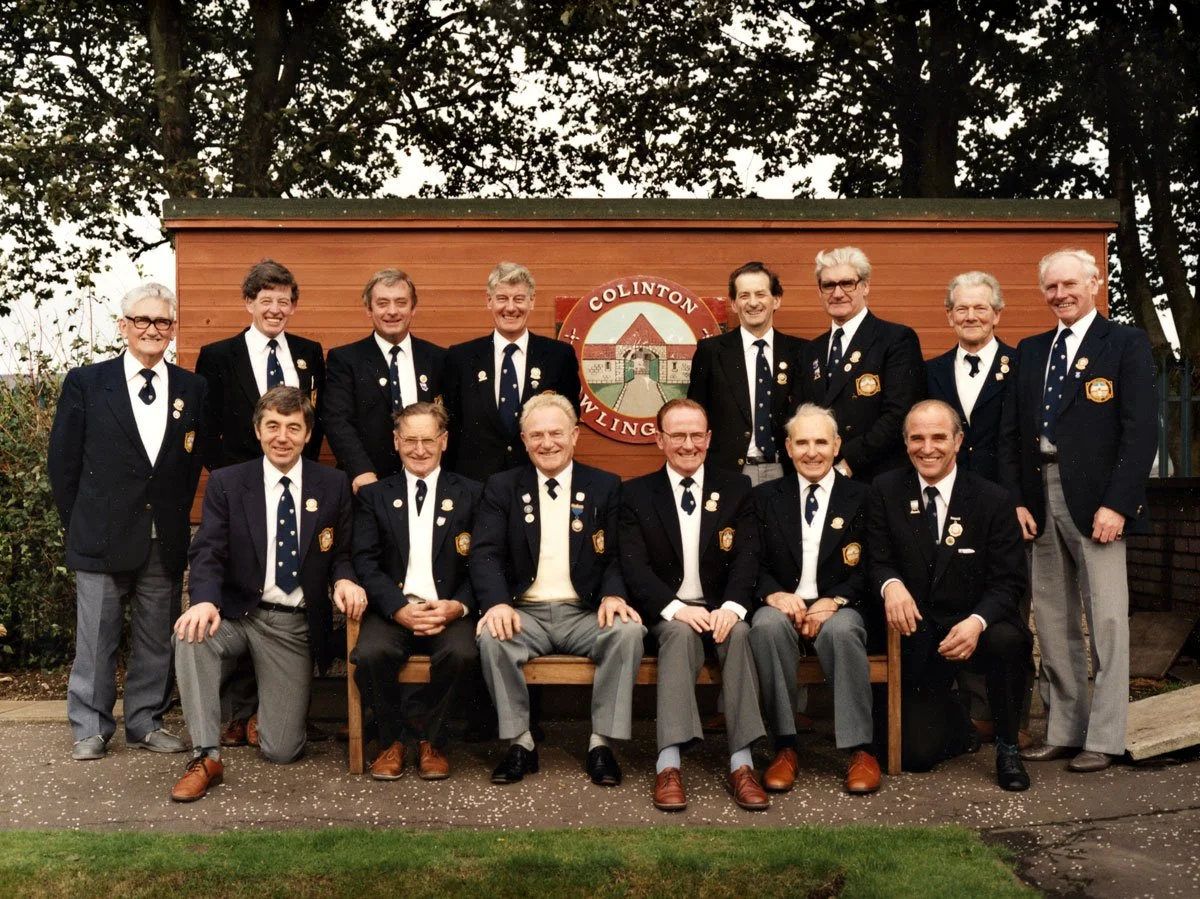 Group of men in formal navy blazers with pins, sitting and standing in front of a wooden building with a circular emblem that reads 'Colinton Waling' and appears to have a horseshoe and landscape scene. Outdoor setting with trees and bushes.