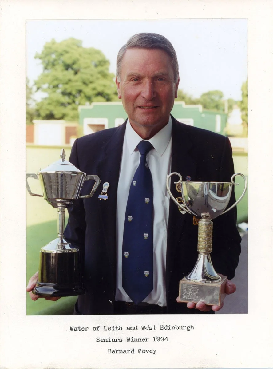 A man in a suit holding two trophies, standing outdoors on a grassy field with trees and buildings in the background.