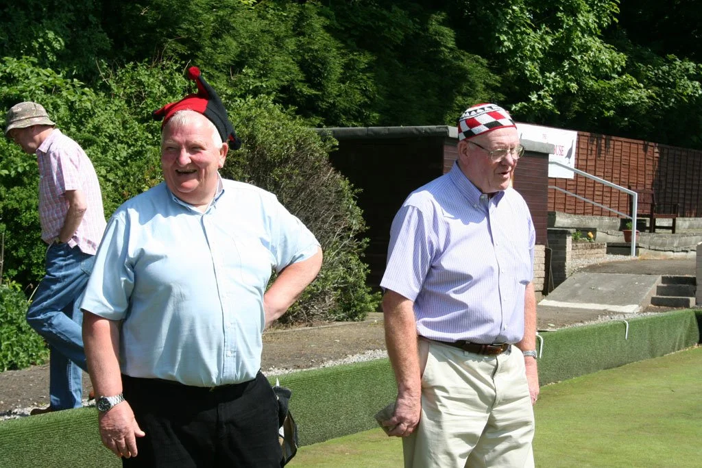 Two men wearing funny hats standing outdoors near a green hedge and steps, with another man in the background.