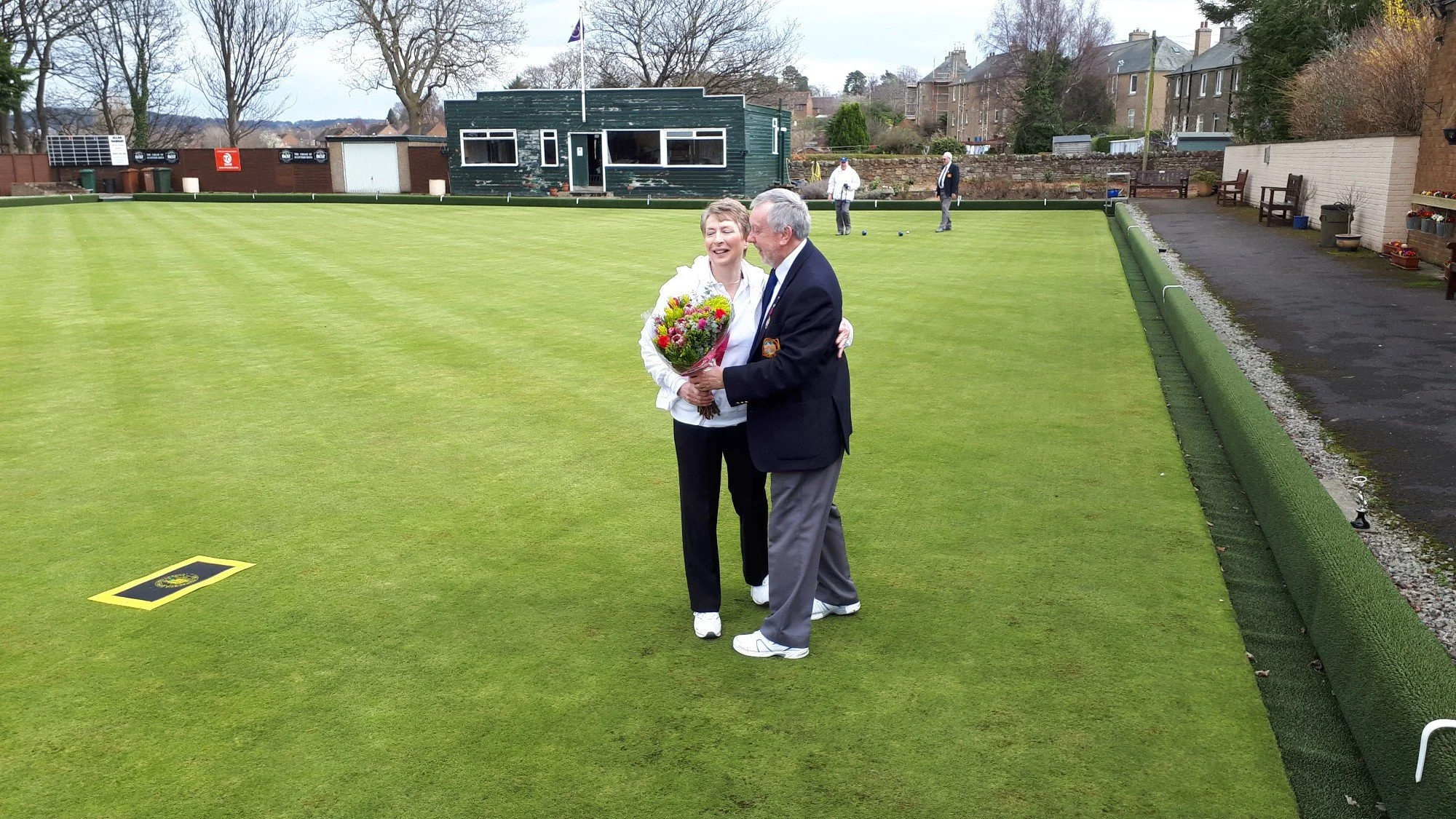 Two people are smiling and hugging on a green lawn, with one holding a bouquet of flowers. In the background, several individuals are playing lawn bowls, with modern houses and trees visible beyond a brick wall.