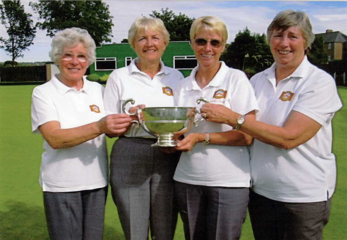 Four women in white shirts standing on a grass field, smiling and holding a large silver trophy together.