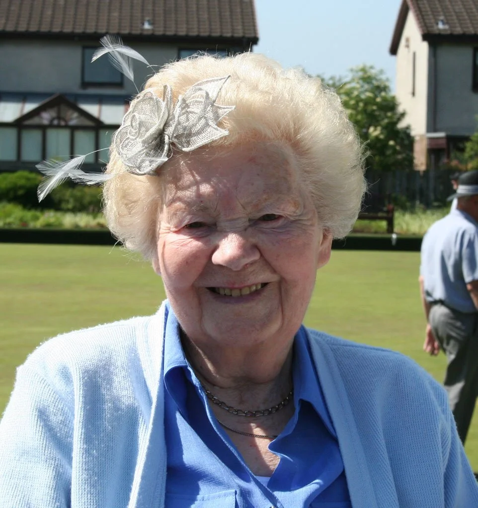 An elderly woman with light blonde curly hair smiling outdoors, wearing a blue shirt, white sweater, and a decorative hair accessory with feathers and a bow.