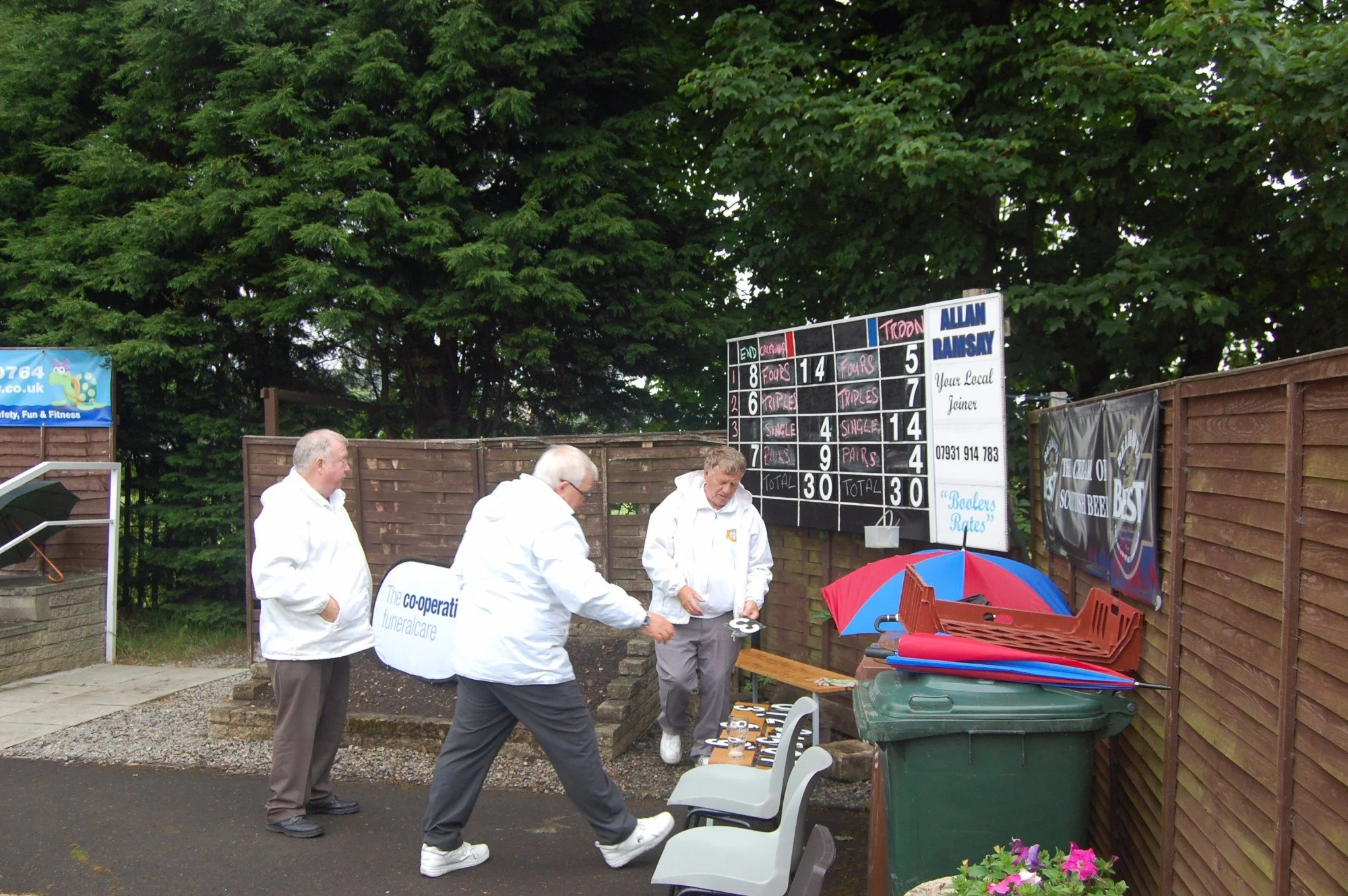 Group of four men in white jackets gathered outside near a board displaying scores, with tables, chairs, umbrellas, and greenery in the background.