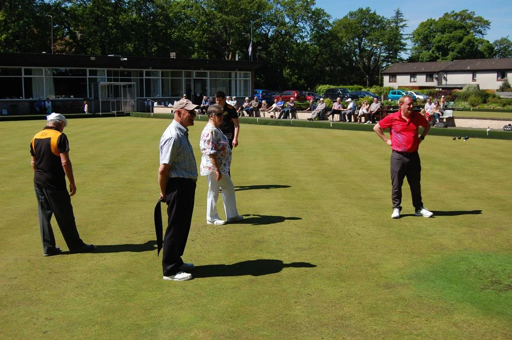 A group of people playing lawn bowls on a green, with spectators sitting on benches in the background and a building nearby.