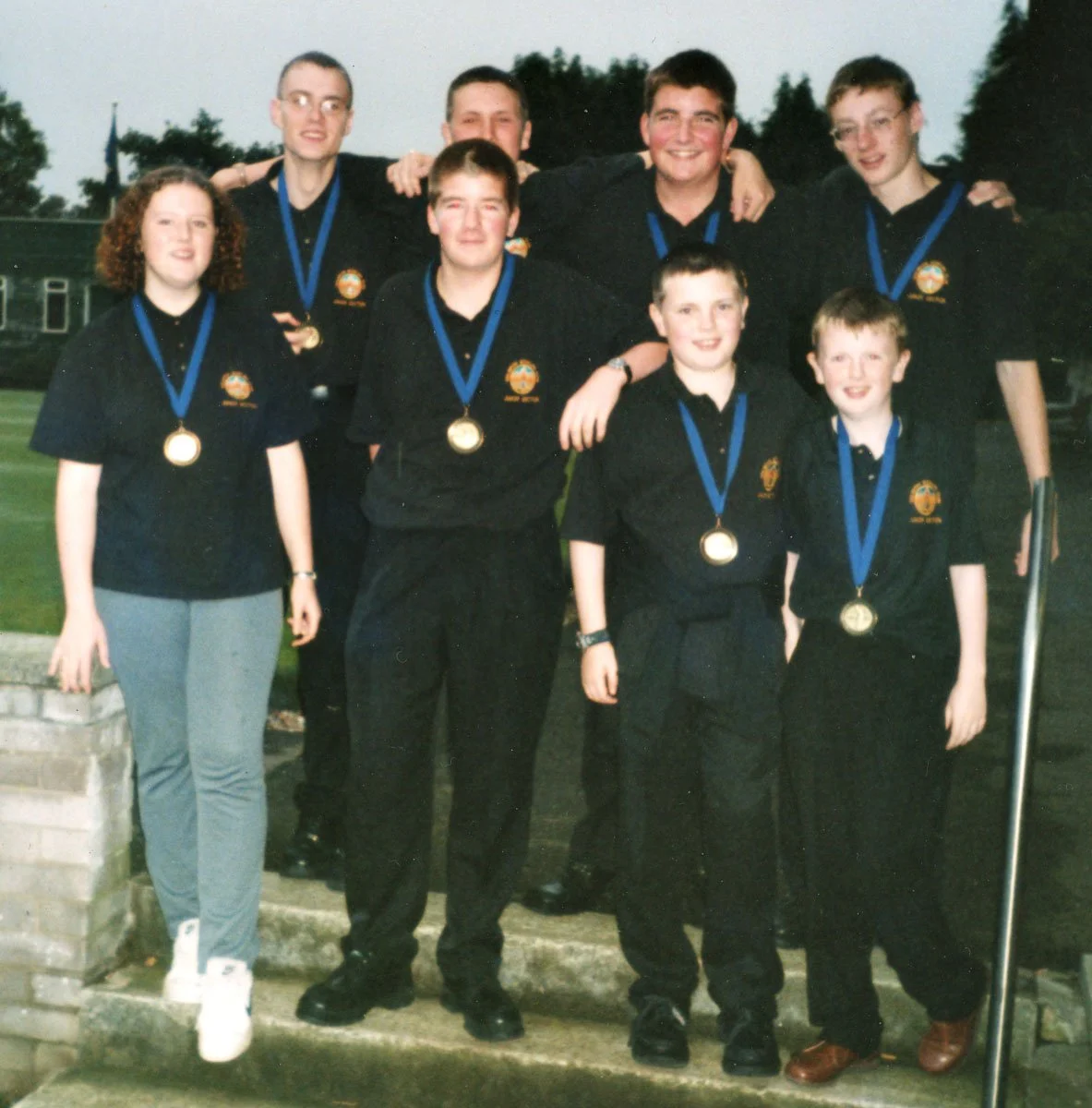 Group of nine young people, seven boys and two girls, wearing black uniforms with medals around their necks, posing outdoors on steps with trees and a building in the background, celebrating their achievement.