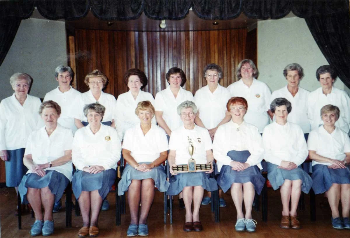 Group of women in white blouses and blue skirts posing for a photo indoors, with one woman holding a trophy. The setting appears to be a stage with wooden paneling and dark curtains.