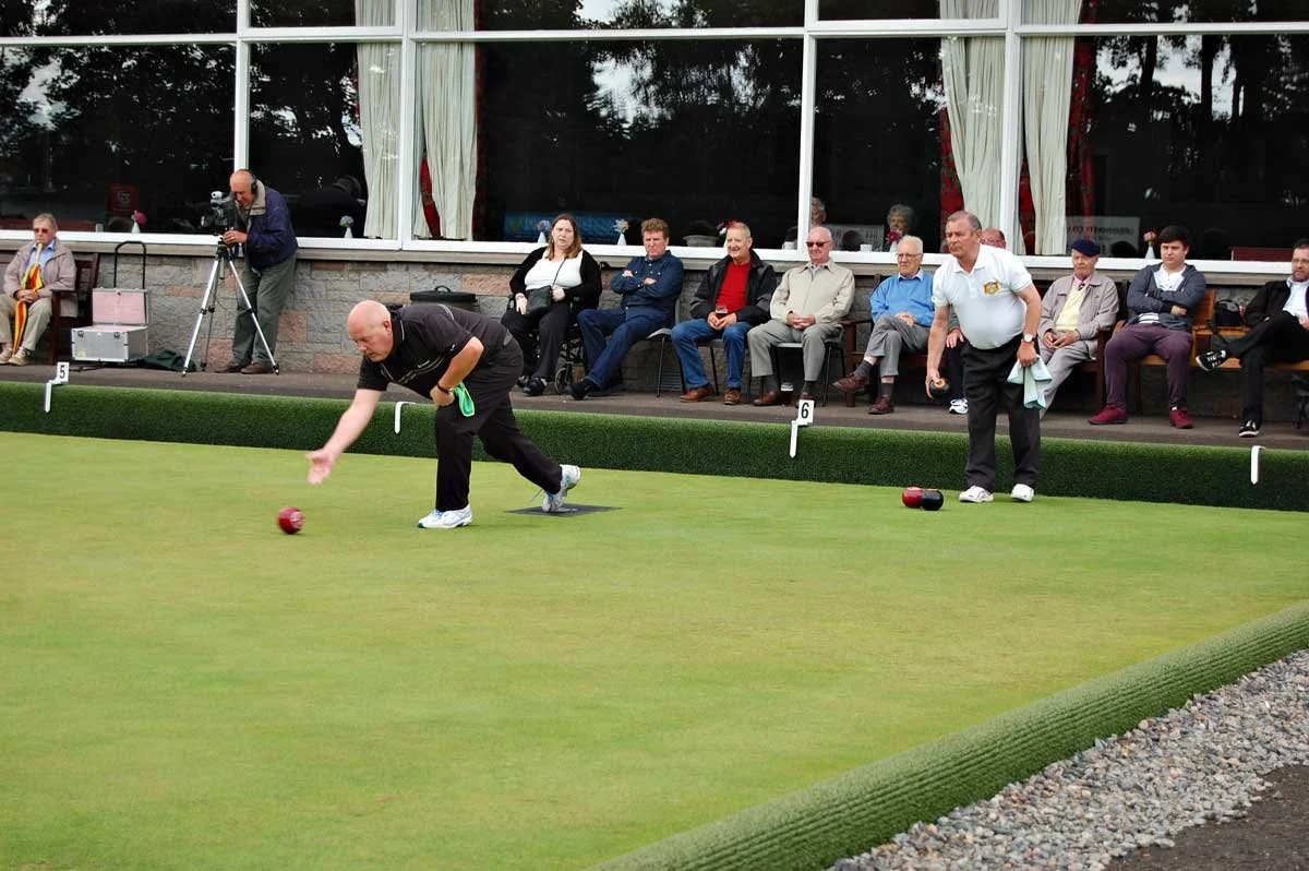 Bocce players on a green field, with spectators seated behind, one player in the process of throwing a bocce ball, others watching, some with cameras, during a game at a recreational or competitive venue.