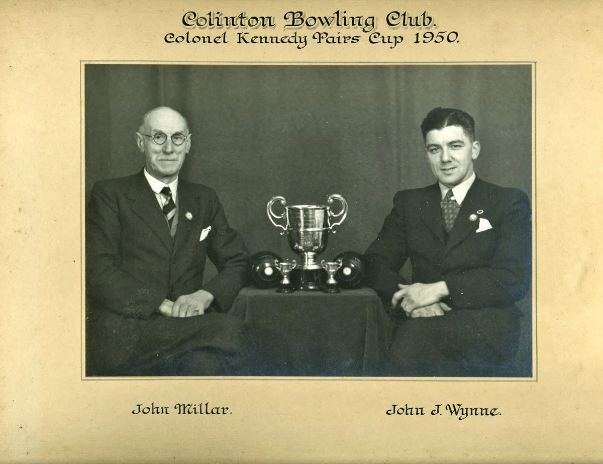 Black and white photograph of two men in suits sitting at a table with a trophy between them. The text above reads "Clifton Bowling Club, Colonel Kennedy Pairs Cup 1950," and the names below are "John Millar" and "John J. Wynne."