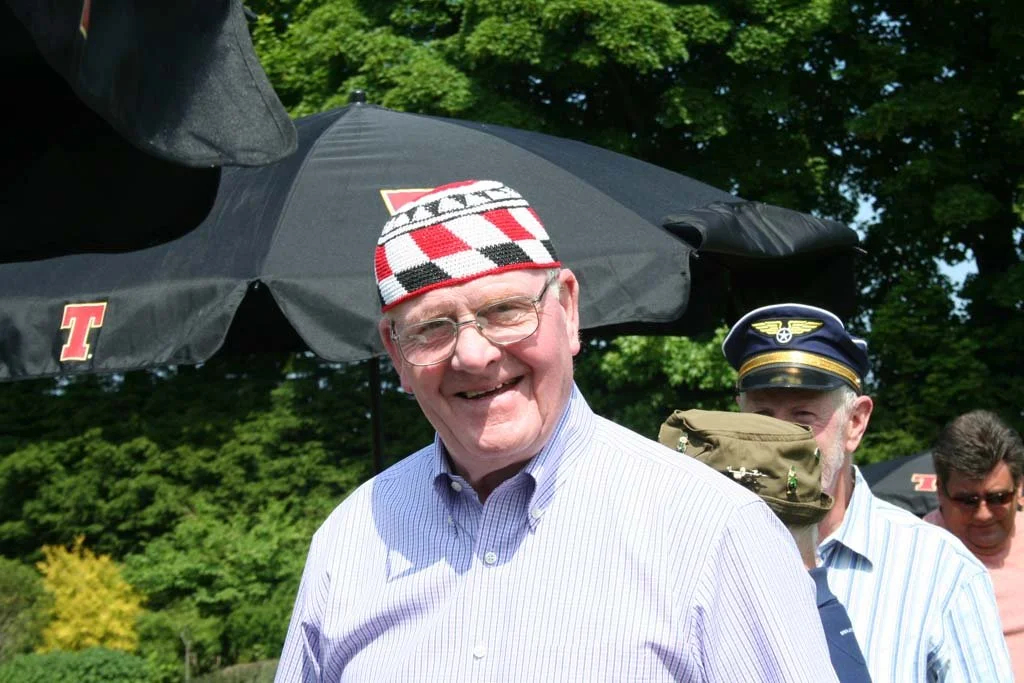 Man smiling, wearing glasses, a striped button-down shirt, and a patterned beanie, outdoors under a large umbrella with trees in the background.