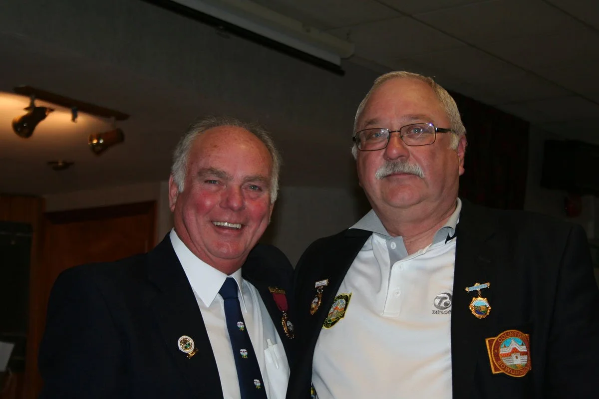 Two men in suits with pins on their jackets, standing close together indoors, smiling at the camera.