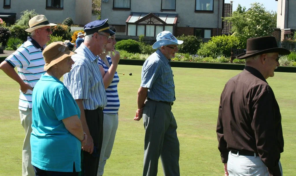 A group of elderly people standing on a lawn, some wearing hats and sunglasses, watching an activity, with buildings and greenery in the background.